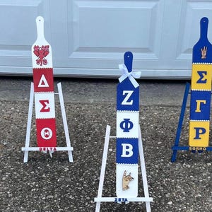 Può includere: Una collezione di pagaie decorative, ognuna con un design unico e lettere greche. Le pagaie sono dipinte in vari colori, tra cui rosa, verde, rosso, blu e nero, e sono esposte su piccoli cavalletti bianchi.