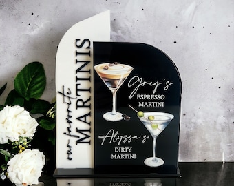 May include: A decorative acrylic sign featuring a white arched panel and a black arched panel. The text reads "our favorite MARTINIS", "Greg's ESPRESSO MARTINI", and "Alyssa's DIRTY MARTINI". Illustrations of an espresso martini and a dirty martini are included.