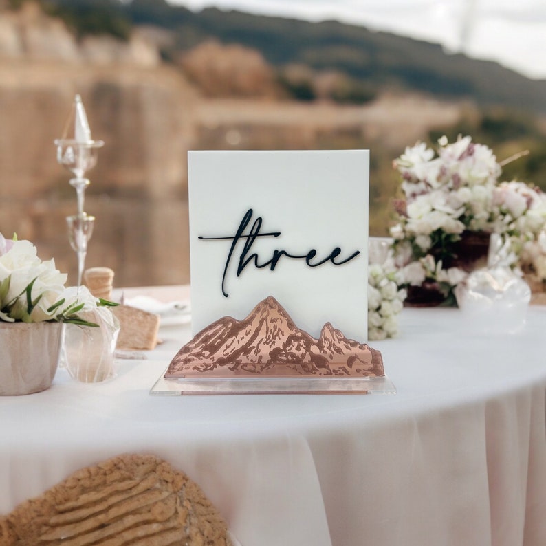 May include: A rectangular white acrylic table sign featuring the word three in black cursive script. The base of the sign is made of clear acrylic with a metallic copper-colored mountain range silhouette attached to the front of the display.