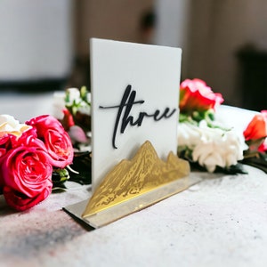 May include: A rectangular white acrylic table sign featuring the word three in black script lettering. The base of the sign is adorned with a gold-colored mountain range silhouette. The sign is positioned upright on a flat surface.