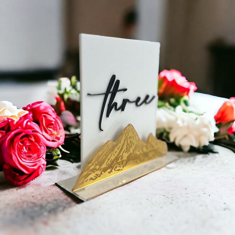 May include: A rectangular white acrylic table sign featuring the word three in black script lettering. The base of the sign is adorned with a gold-colored mountain range silhouette. The sign is positioned upright on a flat surface.
