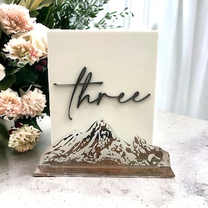 May include: A rectangular off-white table sign featuring the word three in a dark, cursive script. The base of the sign is a clear acrylic stand with a detailed, silver-toned mountain range graphic etched across the front surface.