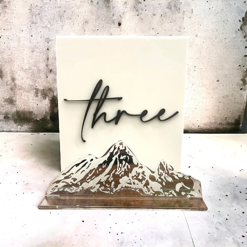May include: A rectangular off-white acrylic sign featuring the word three in black cursive script. Attached to the front is a clear acrylic base with a detailed, etched mountain range graphic that spans the width of the sign.