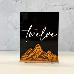 May include: A rectangular black acrylic sign featuring the word twelve in white cursive script. Below the text is a layered mountain range design in a metallic gold finish. The sign is supported by a clear acrylic base stand.