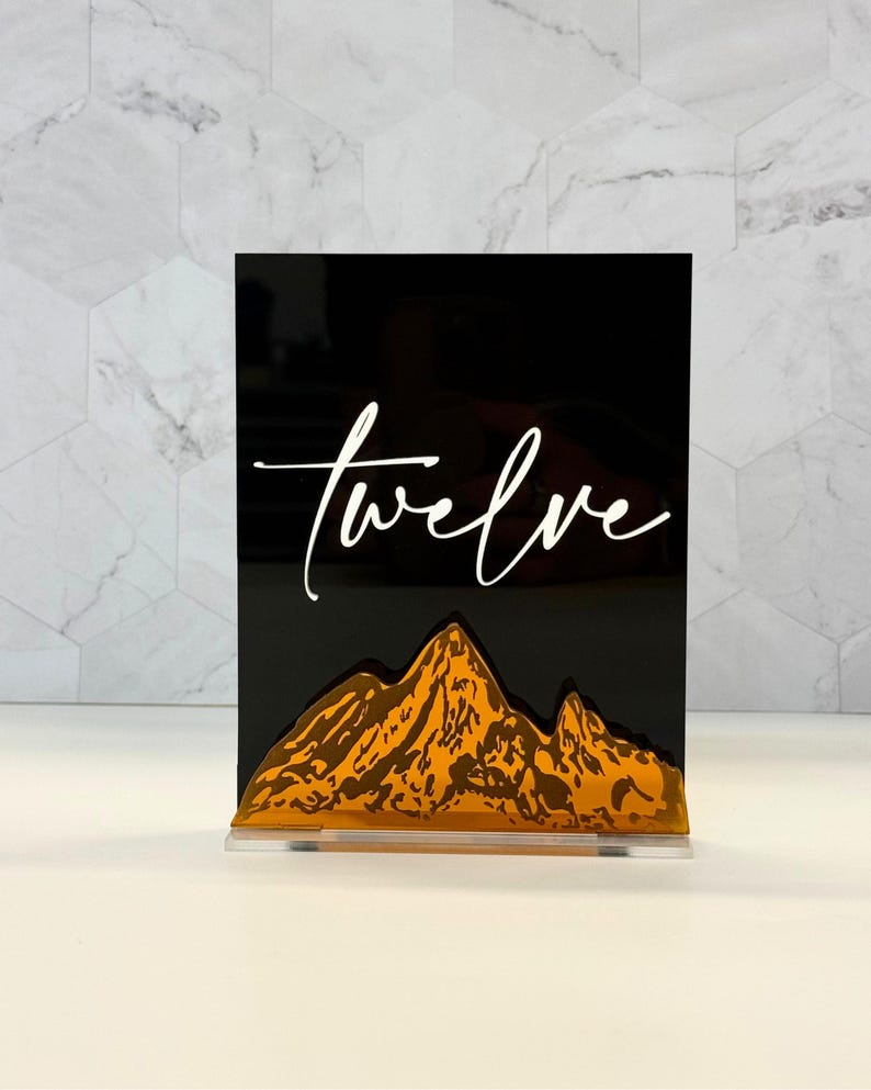 May include: A rectangular black acrylic sign featuring the word twelve in white cursive script. Below the text is a layered mountain range design in a metallic gold finish. The sign is supported by a clear acrylic base stand.