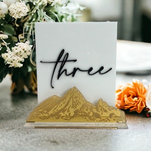May include: A rectangular white acrylic table sign featuring the word three in black cursive script. The base of the sign is decorated with a gold-toned mountain range silhouette that spans the width of the bottom edge of the white panel.