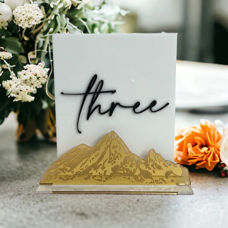 May include: A rectangular white acrylic table sign featuring the word three in black cursive script. The base of the sign is decorated with a gold-toned mountain range silhouette that spans the width of the bottom edge of the white panel.