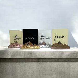May include: Four rectangular acrylic table signs with mountain-shaped bases in metallic finishes. The signs feature the words two, one, three, and four in black script lettering. The backgrounds are light yellow, black, white, and light yellow.