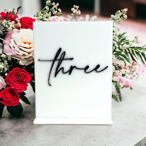 May include: White acrylic table number with the word "three" written in black calligraphy.