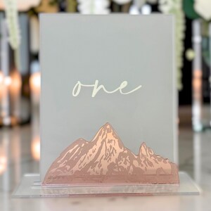 May include: A rectangular frosted acrylic table sign featuring the word one in white cursive script. The base of the sign is decorated with a metallic rose gold mountain range graphic. The entire piece is supported by a clear acrylic stand.