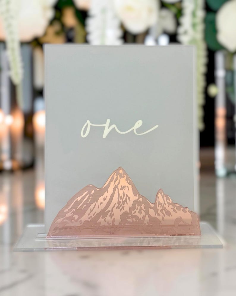 May include: A rectangular frosted acrylic table sign featuring the word one in white cursive script. The base of the sign is decorated with a metallic rose gold mountain range graphic. The entire piece is supported by a clear acrylic stand.