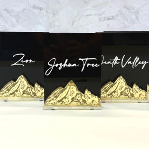 May include: Three rectangular black acrylic signs featuring white cursive text reading Zion, Joshua Tree, and Death Valley. Each sign has a gold-colored mountain range graphic at the base, mounted on a clear acrylic stand for upright display.