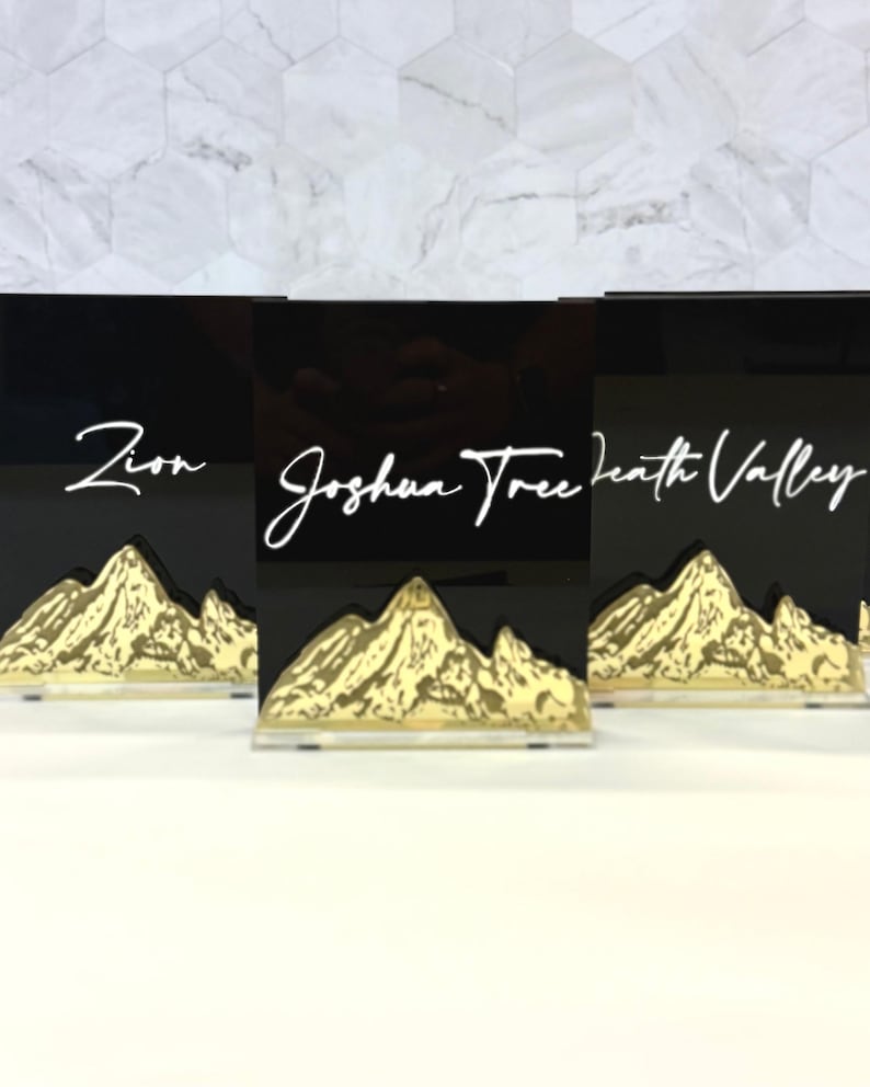 May include: Three rectangular black acrylic signs featuring white cursive text reading Zion, Joshua Tree, and Death Valley. Each sign has a gold-colored mountain range graphic at the base, mounted on a clear acrylic stand for upright display.