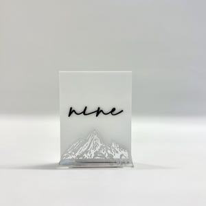 May include: A rectangular white acrylic table sign featuring the word nine in black cursive script. The lower portion of the sign displays a detailed, etched mountain range graphic. The sign is supported by a clear acrylic base stand.