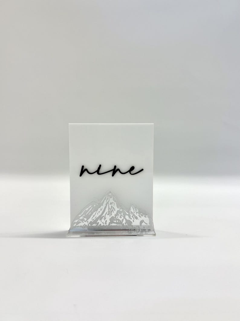 May include: A rectangular white acrylic table sign featuring the word nine in black cursive script. The lower portion of the sign displays a detailed, etched mountain range graphic. The sign is supported by a clear acrylic base stand.
