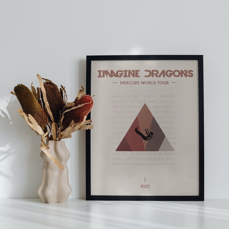 Pode incluir: Um p&ocirc;ster emoldurado em preto com fundo branco e um gr&aacute;fico triangular marrom. O p&ocirc;ster apresenta o texto "Imagine Dragons - Mercury World Tour" e uma lista de t&iacute;tulos de m&uacute;sicas. O ano 2022 est&aacute; impresso na parte inferior do p&ocirc;ster.