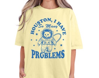 May include: A yellow t-shirt with a blue graphic of a cat wearing an astronaut suit. The text on the shirt reads "Houston, I have so many problems".