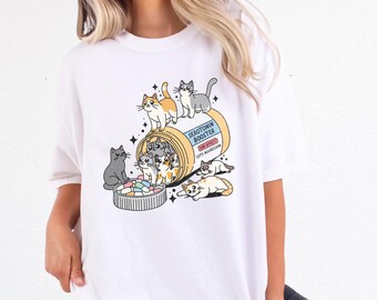 May include: White t-shirt with a graphic of a pill bottle labeled "Serotonin Booster" with cartoon cats spilling out of it. The side effects are listed as "Cute Aggression".
