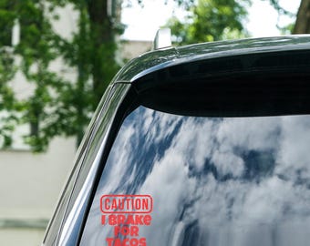 Może przedstawiać: Czerwona naklejka na szybie samochodu z napisem "CAUTION I BRAKE FOR TACOS". Naklejka jest prostokątna z zaokrąglonymi rogami. Szyba samochodu odbija niebo i drzewa.