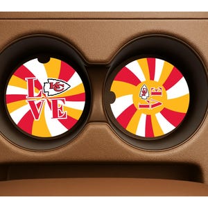 Puede incluir: Dos posavasos para coche con un diseño radial rojo, amarillo y blanco. La palabra "LOVE" se muestra en blanco, con el logotipo de los Kansas City Chiefs. Los posavasos están en los portavasos de un coche.