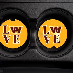 May include: Two car coasters with a gold background and the letters "LW VE" in burgundy and white. The coasters are in a black car cup holder.