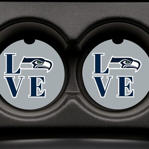 Może przedstawiać: Dwa podkładki do samochodu z szarym tłem i napisem "LOVE" w granatowych literach. "O" zastąpiono logo Seattle Seahawks w kolorze granatowym i białym. Podkładki znajdują się w czarnym uchwycie na kubek w samochodzie.