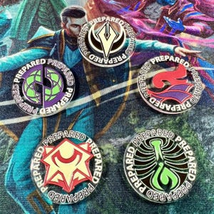 Puede incluir: Una colección de cinco pines redondos de metal con varios diseños coloridos. Cada pin tiene la palabra "PREPARED" alrededor del borde. Los diseños incluyen un emblema estilizado, un globo, una llama, una estrella y un esqueleto.