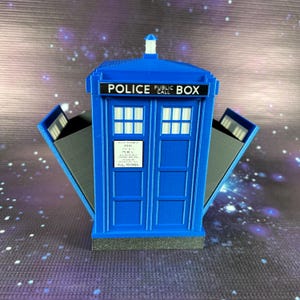 May include: A blue and gray, 3D printed, TARDIS-style box with a sign that reads "Police Public Call" and a small, white light on top. The doors are open, revealing the interior.