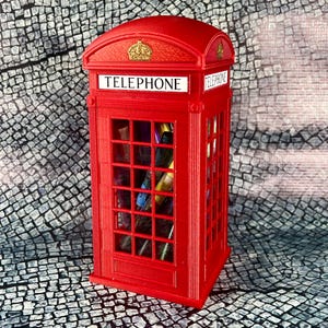 May include: Red telephone booth shaped pencil holder with a crown on top and the word "TELEPHONE" on the front. The holder is filled with colorful pens and pencils.
