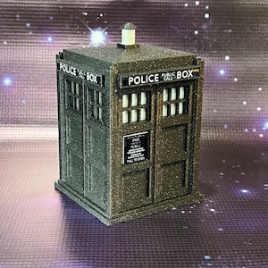 May include: A dark gray, textured, miniature replica of a police call box with the words "Police Public Call Box" printed on the front. The box has a small sign that reads "Free Parking For TARDIS Only.  All Other Vehicles Will Be Towed Away.  Pull To Open."