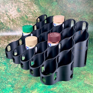 May include: Black 3D-printed paint bottle organizer. The organizer has multiple curved slots designed to hold small paint bottles. Several bottles of paint are visible, with colors including green, tan, and brown. The organizer is on a green and brown surface.