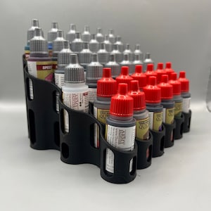May include: Black plastic paint bottle holder with rows of slots for holding bottles of paint. The holder is filled with bottles of paint with silver and red caps. The bottles have labels that say "Army Painter" and "The Army Painter" and "The Army Painter Quick Shade".