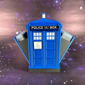 May include: A blue and black 3D printed TARDIS card holder with the words "POLICE PUBLIC CALL" on the front. The doors are open and the interior is visible.