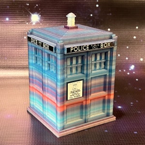 May include: A blue and pink police box with a white roof and a sign that says "Police Public Call Box". The box is made of plastic and has a glittery finish.