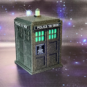 May include: A dark gray, textured, miniature replica of a police call box with a light on top. The box has a sign that reads "FREE PUBLIC CALL" and "PULL TO OPEN".
