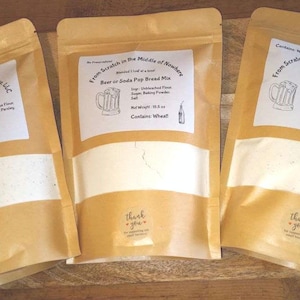 May include: Three brown paper bags with white labels. Each bag contains a beer bread mix. The labels have the text "From Scratch in the Middle of Nowhere, LLC" and list the ingredients and net weight of the mix. The labels also have the text "Contains Wheat".