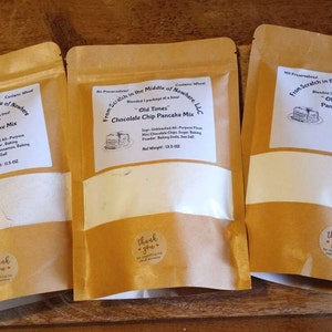 May include: Three brown paper bags with white labels. Each bag contains a pancake mix. The labels have the text "From Scratch in the Middle of Nowhere" and "Old Times" with different pancake mix flavors: Cinnamon, Chocolate Chip, and plain. Each bag has a "Thank you" sticker.