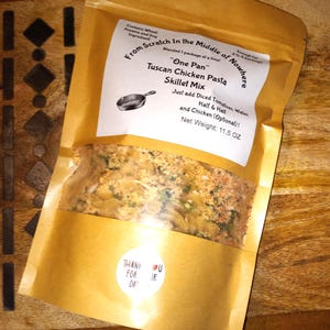 May include: A brown paper bag with a white label that reads "From Scratch In the Middle of Nowhere" and "Tuscan Chicken Pasta Skillet Mix". The label also includes the text "One Pan" and "Net Weight: 11.5 OZ". The bag contains a mixture of dried pasta, herbs, and spices.