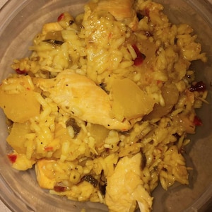 K&ouml;nnte beinhalten: Eine Nahaufnahme einer Sch&uuml;ssel mit gelbem Reis mit H&uuml;hnchen und Ananas. Der Reis ist gekocht und fluffig, und das H&uuml;hnchen ist zart und saftig. Die Ananas ist s&uuml;&szlig; und s&auml;uerlich, und das Gericht ist mit roten Pfefferflocken garniert.