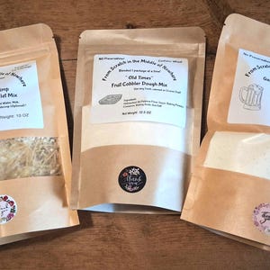 May include: Three kraft paper pouches of dry food mixes. The pouches have labels with text and illustrations. One is for Alfredo pasta, another for fruit cobbler, and the third for garlic beer bread. Each pouch has a circular sticker with floral accents.