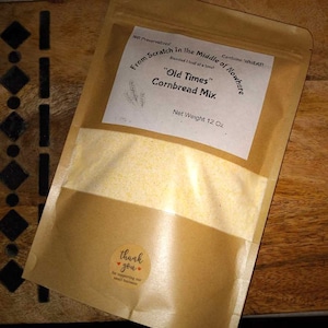 May include: A brown paper bag with a white label that reads "From Scratch in the Middle of Nowhere" and "Old Times Cornbread Mix." The bag contains a yellow cornbread mix and has a sticker that says "thank you" for supporting small businesses. The bag is 12 ounces.