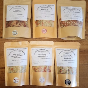 May include: Six brown paper pouches with clear plastic windows, each containing a different type of soup mix. The pouches are labeled with the text "From Scratch in the Middle of Nowhere" and the name of the soup. The soups include "Unstuffed Bell Pepper Soup", "Warm You Up Chicken & Rice Soup", "Hearty Barley Vegetable Tomato Soup", "Warm You Up Hearty Vegetable Beef with Rice Soup", "Quick Warm You Up Chicken Noodle Soup", and "Quick Warm You Up Hearty Vegetable Beef Noodle Soup".