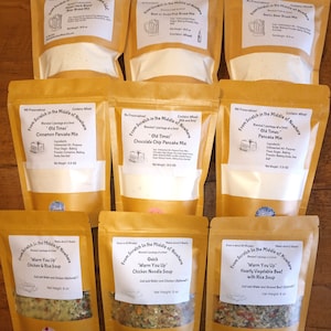 May include: Nine brown paper bags with white labels containing different food mixes. The labels have the text "From Scratch in the Middle of Nowhere" and list the ingredients and net weight of each mix. The mixes include "Old Times" Cinnamon Pancake Mix, "Old Times" Chocolate Chip Pancake Mix, "Old Times" Pancake Mix, "Warm You Up" Chicken & Rice Soup, "Warm You Up" Quick Chicken Noodle Soup, and "Warm You Up" Hearty Vegetable Beef with Rice Soup.