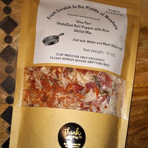 May include: A brown paper bag with a white label that reads "From Scratch In the Middle of Nowhere" and "Unstuffed Bell Pepper with Rice Skillet Mix". The bag contains a mixture of rice and red bell peppers. The label also states "Net Weight: 10 OZ" and "CLAY MOISTURE PACK ENCLOSED! PLEASE REMOVE BEFORE EMPTYING BAG!"