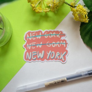 May include: A pink sticker with the words "NEW YORK" written in a stylized font. The words "NEW GORK" and "NEW GORM" are crossed out above the correct spelling.