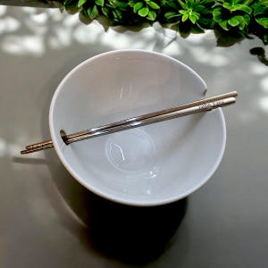 May include: A white ceramic bowl with a pair of silver chopsticks resting inside. The chopsticks are engraved with the name "Justin Yang".