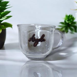 May include: A clear glass mug with a double wall design. The mug has a handle and the name "Angela" is written on the side in brown lettering.