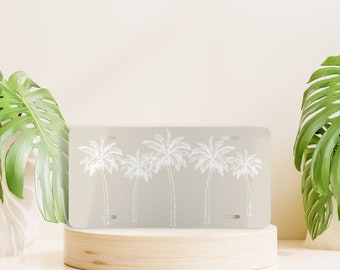 May include: A white metal license plate with a design of four palm trees in a row. The license plate is on a light wood surface.