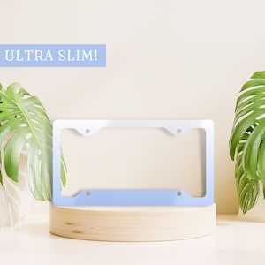 May include: A white and light blue license plate frame with the text "ULTRA SLIM!" on a light beige background. The frame is sitting on a wooden platform.