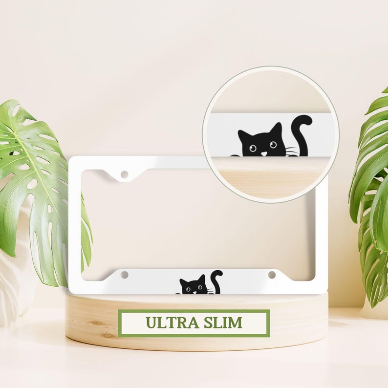 May include: White metal license plate frame with a black cat silhouette peeking over the top. The frame is labeled "ULTRA SLIM".
